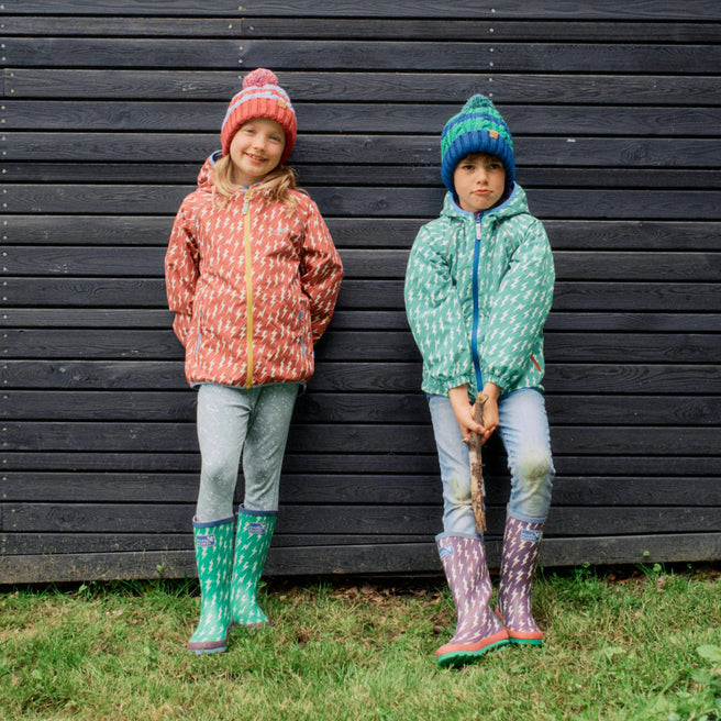 Two children wearing hats and patterned raincoats, standing against a black wooden wall in green boots and trousers in front of grass.